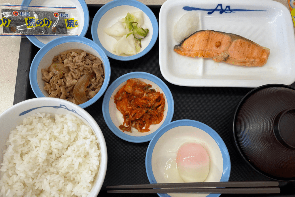 This image shows a traditional Japanese meal served on a tray, featuring a variety of dishes. The meal includes a bowl of steamed white rice, a serving of grilled salmon with grated radish, a bowl of simmered beef and onions, kimchi, pickled vegetables, a soft-boiled egg, miso soup, and a packet of nori (seaweed). The meal is neatly arranged, with chopsticks placed in front of the soft-boiled egg bowl, ready to eat.