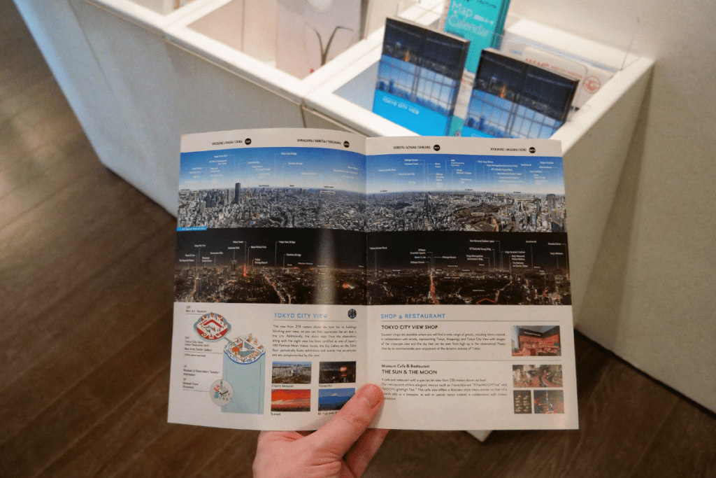 The image shows a hand holding a folded paper guide that provides a panoramic view of Tokyo, with labels marking famous landmarks. The top half of the guide features daytime and nighttime views of the cityscape, with various buildings and locations identified. The bottom section includes information about the Tokyo City View observation deck, a shop, and a restaurant called "The Sun & The Moon," along with illustrations of featured attractions like cherry blossoms, fireworks, and sunset views. Brochures are also visible in the background.