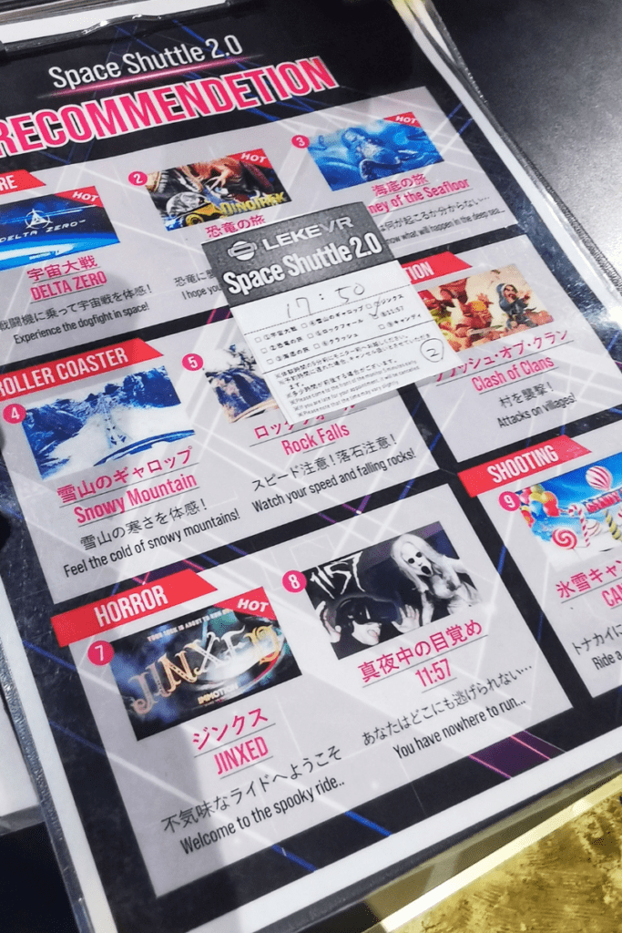 The image features a promotional display of various virtual reality experiences available at a venue, titled "Space Shuttle 2.0 RECOMMENDATION." The display includes colorful posters and descriptions for different VR themes such as "Delta Zero" for a space dogfight experience, "Journey of the Sea" featuring underwater adventures, "Snowy Mountain" for a cold mountainous environment, "Rock Falls" involving high-speed action and falling rocks, and "Jinxed" which is a horror-themed ride. Each poster includes vivid graphics, thematic descriptions, and is marked with labels like "HOT" to indicate popular choices.