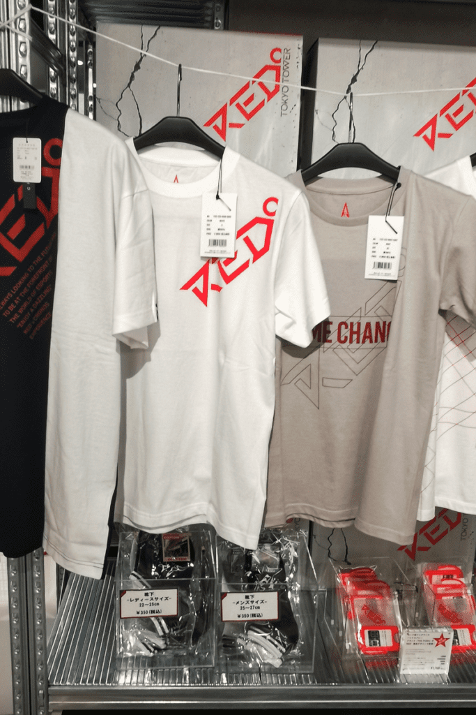 The image shows a display of stylish t-shirts for sale at a store, featuring official "Red" merchandise. The shirts come in various colors such as white and beige, with bold red and gray graphic designs. Each shirt has a tag attached, and the shelves below hold additional items, including socks and other accessories packaged in plastic. The branding and design give a modern, minimalist vibe.