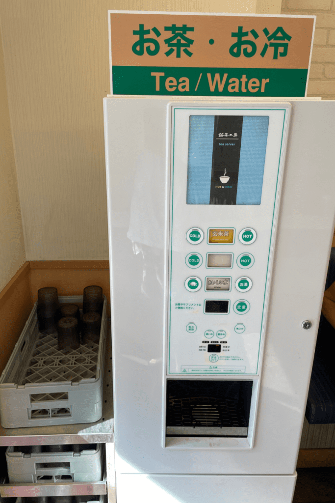 This image shows a self-service tea and water dispenser at a Matsuya restaurant. The machine offers both hot and cold options, with buttons for dispensing cold water, hot water, and different types of tea, including genmai (brown rice) tea. A stack of reusable plastic cups is stored in crates to the left of the machine, allowing customers to serve themselves. The signage at the top is labeled "Tea / Water" in both Japanese and English.