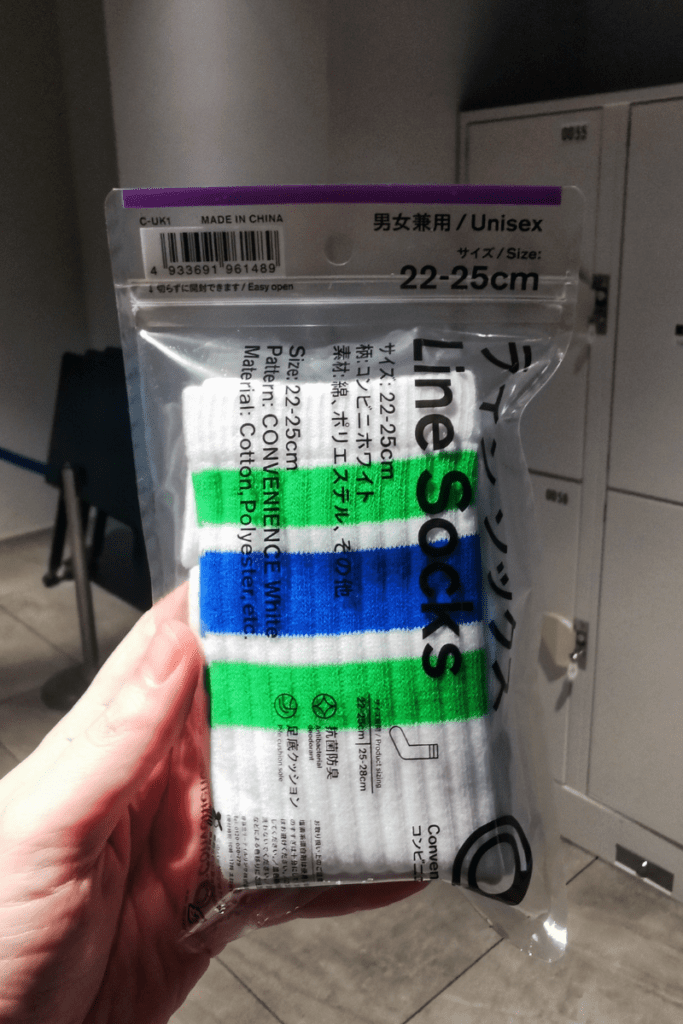 The image shows a packaged pair of "Line Socks" being held up, labeled as unisex and sized 22-25 cm. The socks feature a striped design in white, blue, and green, and the packaging provides details about the materials, which are cotton and polyester. The packaging also includes instructions for easy opening and product care symbols. In the background, lockers are visible, indicating a facility where visitors can store their belongings. The image suggests that socks may be required for certain activities, with lockers conveniently available nearby.
