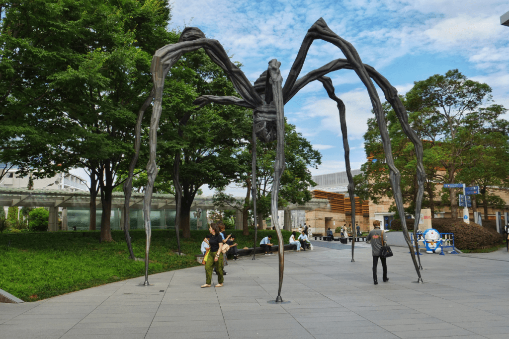 The image shows the "Maman" spider sculpture, a large bronze structure with long, spindly legs, situated in an outdoor plaza. The sculpture is surrounded by trees and greenery, with several people walking nearby and others sitting on benches. The sky is partly cloudy, and the area appears to be a popular meeting point. The surrounding architecture and open space create a peaceful atmosphere for visitors.