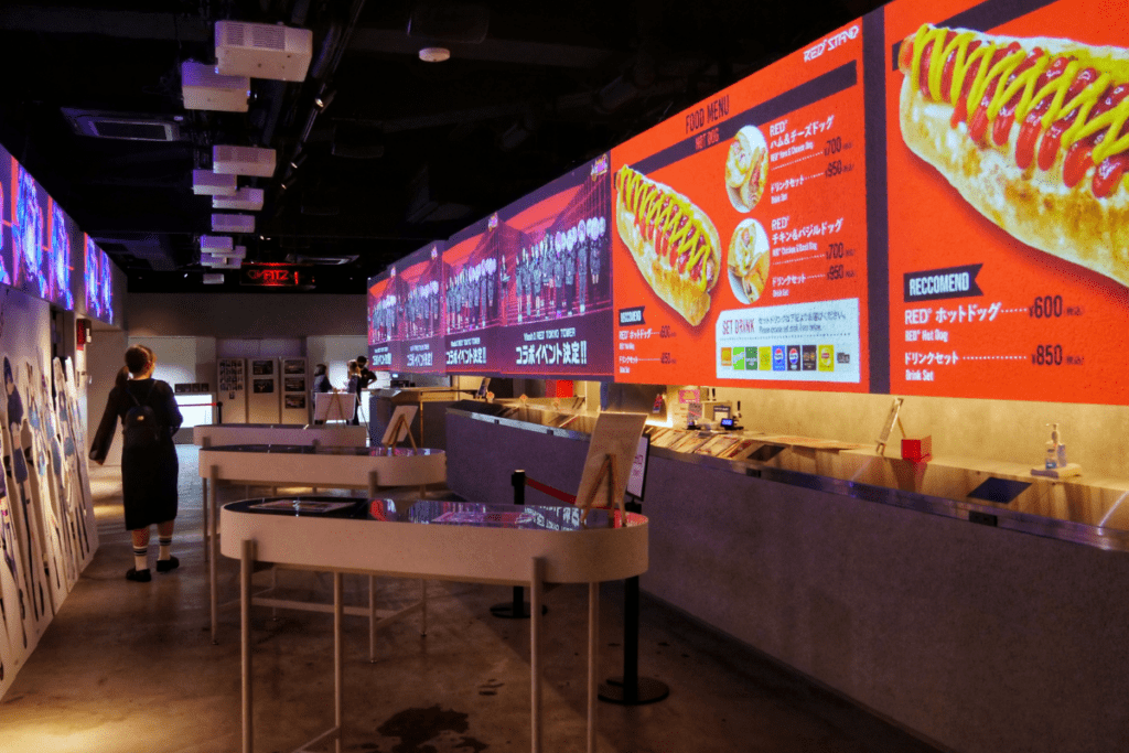 The image shows the interior of "The Red Stand," a food counter offering a variety of food and drinks. Large digital menu boards dominate the space, featuring items like hot dogs covered in mustard and ketchup, along with other snacks such as ham and cheese dogs and churros. The bright orange menus display prices and food descriptions, with combo meal options highlighted. In the foreground, a few tables are scattered around, and a customer is seen walking past cardboard cutouts. The atmosphere is modern and casual, with digital signage and colorful displays enhancing the space.