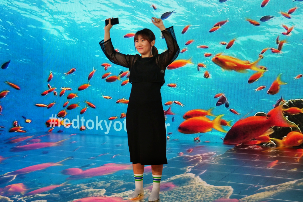 The image shows a young woman posing joyfully with her hands raised in front of a large digital screen displaying an underwater scene filled with colorful fish. She is standing on a reflective floor, and the vibrant oceanic backdrop creates an immersive effect. The hashtag "#redtokyotower" is visible on the screen, indicating the location. The scene is part of a photo opportunity in the Sky Stadium, designed to provide a fun and interactive experience for visitors. The woman is wearing striped socks, adding to the playful atmosphere.