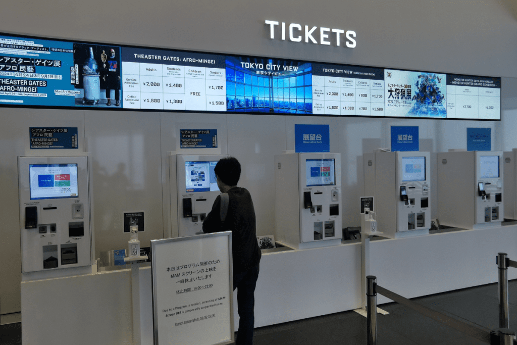 The image shows a ticketing area with several self-service kiosks where people can purchase tickets for various attractions, including Tokyo City View and special exhibitions like "Theaster Gates: Afro-Mingei" and "Monster Hunter 20th Anniversary Exhibition." Above the kiosks, digital displays list ticket prices for adults, students, children, and seniors, with options for both on-site and online admission. A person is seen interacting with one of the kiosks, and signs indicate that the ticketing machines are available in multiple languages, including English.