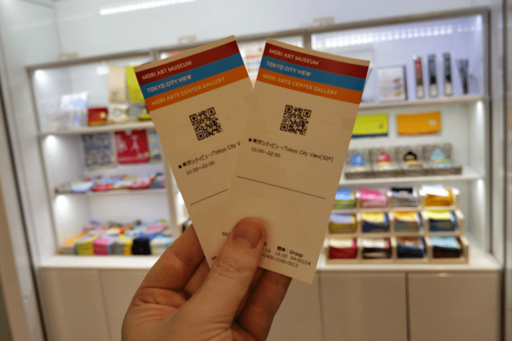 The image shows two tickets being held up, featuring a QR code for entry. The tickets are for the Tokyo City View, located on the 52nd floor, and also include access to the Mori Art Museum and the Mori Arts Center Gallery. In the background, a well-lit gift shop displays colorful souvenirs and products on shelves. The tickets display the hours of operation from 10:00 to 22:00.