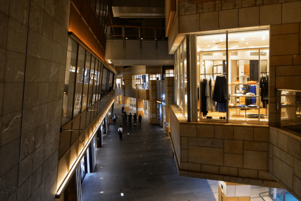 The image shows a modern shopping area, likely part of a complex, with multiple levels of shops and restaurants. One of the stores, visible through large glass windows, displays racks of clothing under bright lighting. The walkway below is lined with storefronts and illuminated by warm, ambient lighting. A few people are walking along the wide, clean corridor, which has a sleek, polished look with stone walls and contemporary architecture. The area exudes a calm, upscale atmosphere for shoppers and diners.