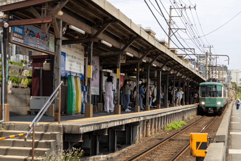 The platform at Kamakurakōkō-Mae Station is bustling with people, as a green Enoshima Electric Railway train approaches on the tracks. The wooden station structure has a rustic look, with overhead wires extending above, and the station’s iconic setting is framed by a mix of local signs and greenery.