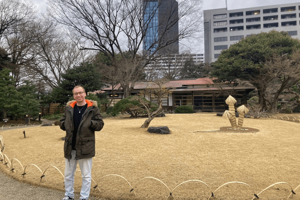 A person wearing a green parka stands smiling in the Koishikawa Korakuen Garden, Tokyo, on a winter day. The garden features neatly trimmed shrubs, traditional buildings in the background, and straw-wrapped trees for winter protection. Tall modern buildings rise in the distant background, contrasting with the natural setting.