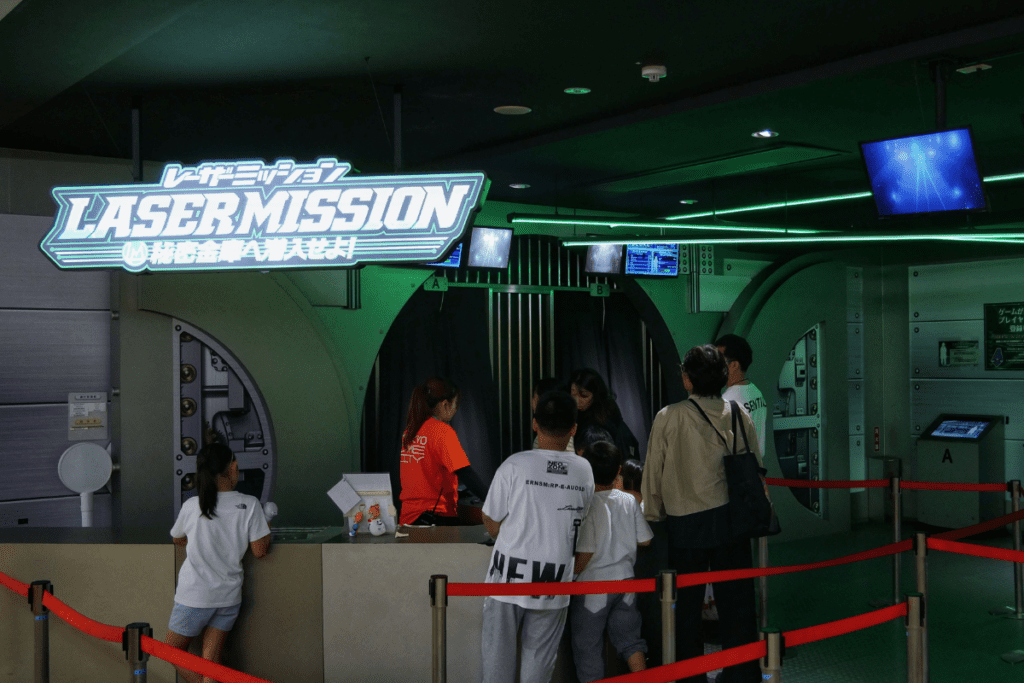 The image shows the entrance to the "Laser Mission" attraction at Tokyo Dome City, featuring a neon-lit sign above a futuristic-themed entryway. Visitors are gathered at the check-in counter, with red ropes guiding the queue. Staff members assist participants as they prepare to enter the mission area, which is designed with metallic accents and glowing green lighting.