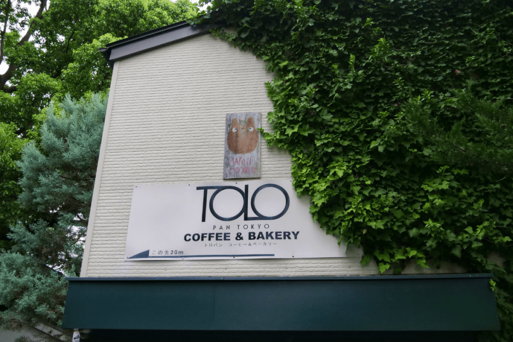 A sign for "TOLO Pan Tokyo Coffee & Bakery" is mounted on the side of a building partially covered with green ivy. Above the sign, a small, wooden plaque featuring a Totoro character with the word "Welcome" adds a whimsical touch. The greenery and Totoro artwork create a cozy and inviting atmosphere for visitors.