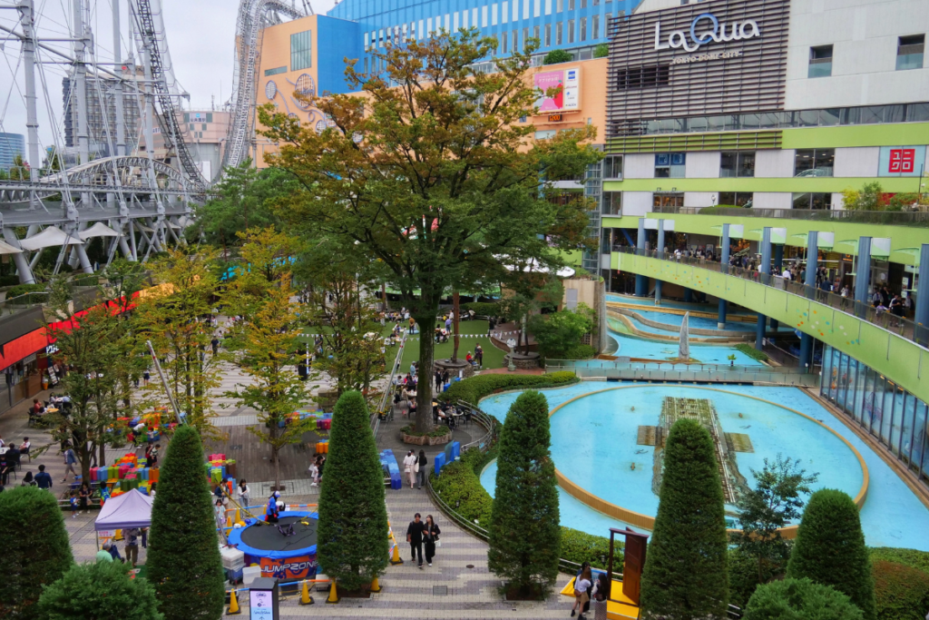 A vibrant scene of the LaQua area at Tokyo Dome City, featuring a lush garden with trees, a winding water feature, and a playground with colorful structures. The LaQua shopping complex is visible in the background, along with a section of the Thunder Dolphin roller coaster's track looping through the building facade. Visitors are scattered throughout the area, enjoying the recreational space.