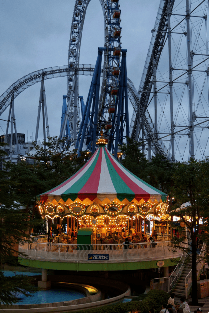 A brightly lit carousel, the Venus Lagoon, at Tokyo Dome City's LaQua, glowing with warm lights against a dusk sky. Behind it, the towering Thunder Dolphin roller coaster tracks create a dramatic backdrop. The colorful carousel contrasts with the steel structure, making the scene lively and enchanting in the evening.