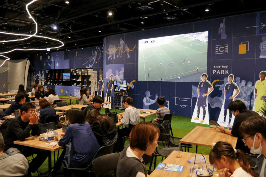 The image shows visitors seated at tables enjoying food and drinks inside the Blue-ing! Cafe & Bar. In the background, a large screen displays a live soccer match, while cutouts of Japanese soccer players decorate the space. The venue's interior features a blue theme with sports-related graphics, creating a lively and sporty atmosphere.