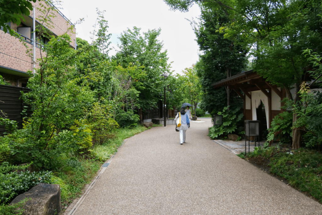 This image shows a peaceful, tree-lined path in a quiet Japanese neighborhood, with lush greenery on both sides. A person holding an umbrella is walking down the path, which curves gently ahead. The serene setting is enhanced by the natural surroundings and the simple, well-maintained architecture of nearby buildings. The scene captures a tranquil moment, offering a glimpse into the calm and nature-filled environment of the Daita area.