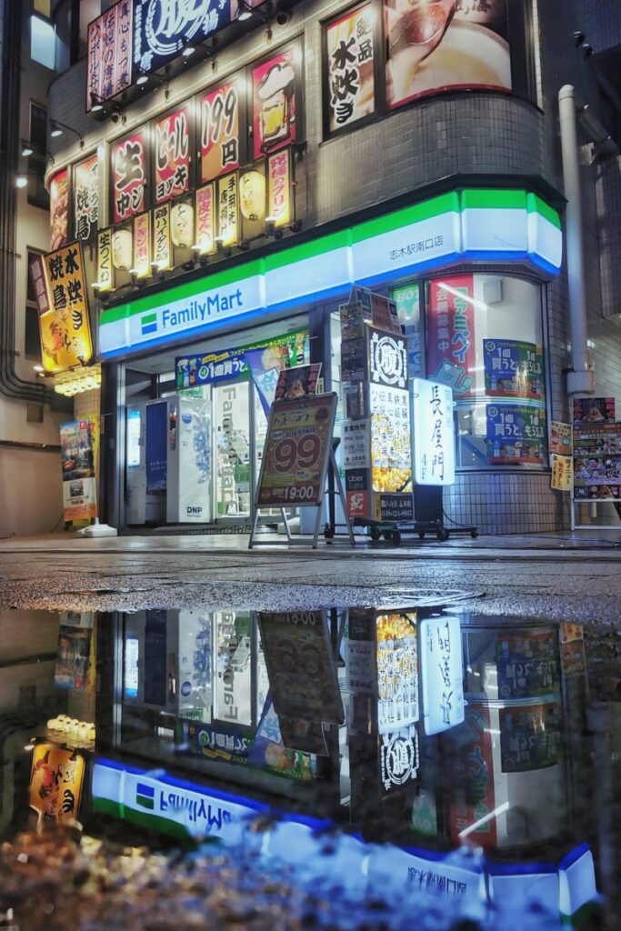 Nighttime photo of a Family Mart storefront in Japan, illuminated with bright lights and colorful advertisements for food and drinks. The wet pavement in front reflects the vibrant neon signage and storefront details, creating a mirrored effect that enhances the urban atmosphere.