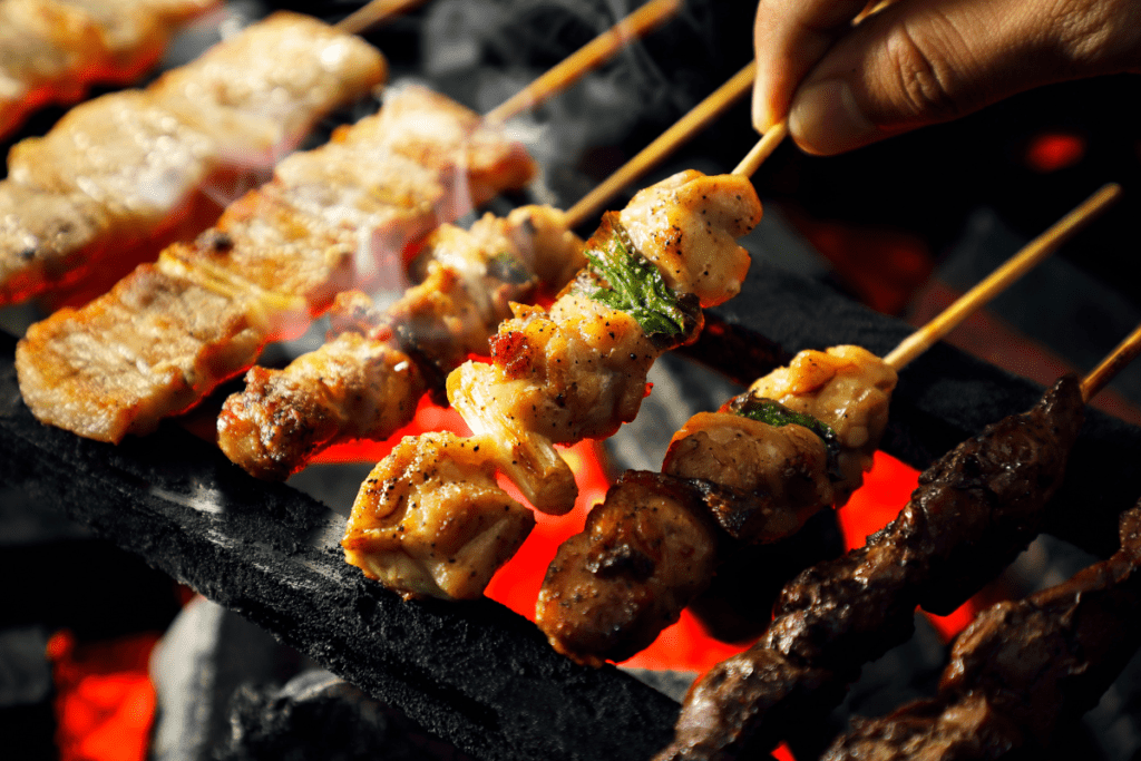 Close-up of skewers of grilled yakitori, including various cuts of seasoned chicken, sizzling over hot coals. Knowing Japanese phrases for restaurants could be helpful for ordering dishes like this, especially to specify preferences or ask about ingredients.