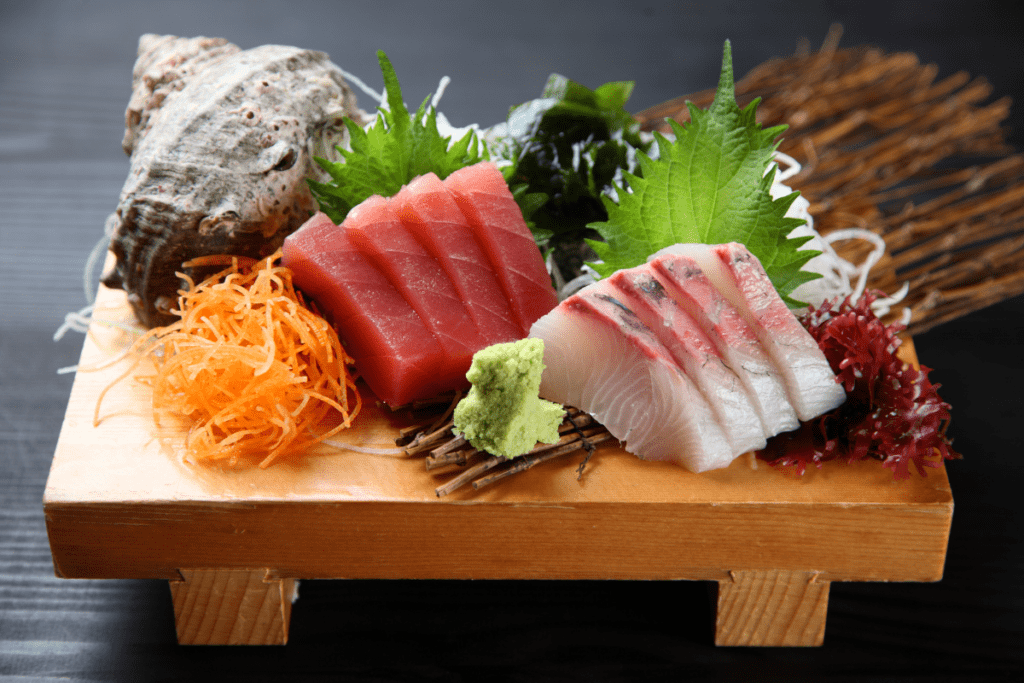 A beautifully presented sashimi platter on a wooden board, featuring slices of fresh tuna and white fish alongside garnishes of shredded carrots, green shiso leaves, and a small mound of wasabi. The arrangement includes a decorative shell and seaweed, highlighting the freshness of the seafood.