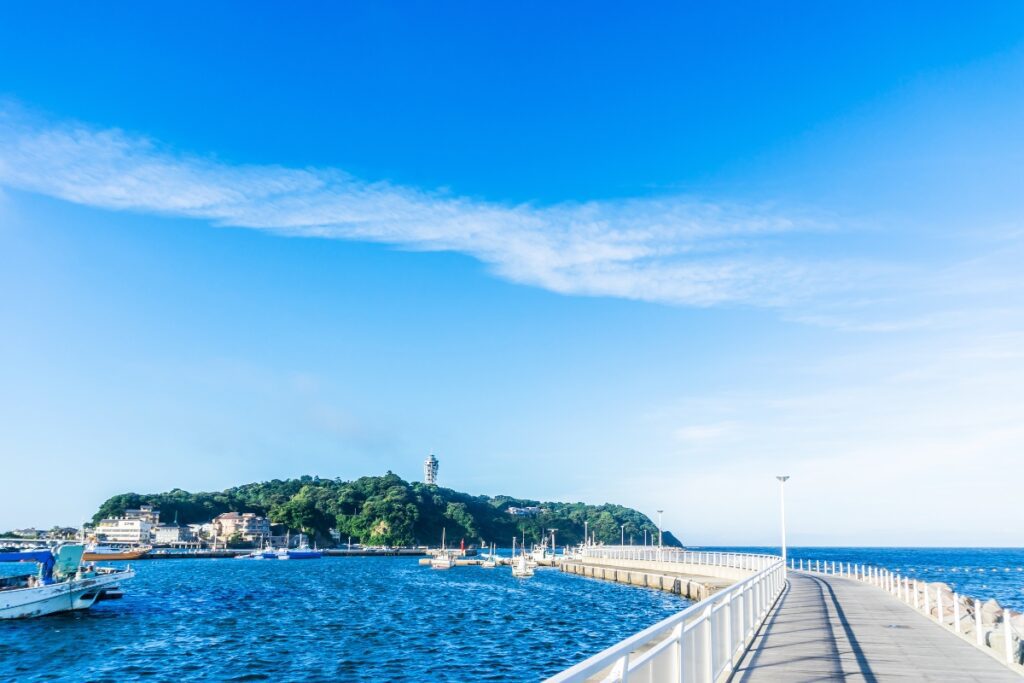 A view of Enoshima Island with its lush greenery and observation tower, surrounded by the ocean. A bridge extends across the water, connecting the island to the mainland, with boats docked nearby.