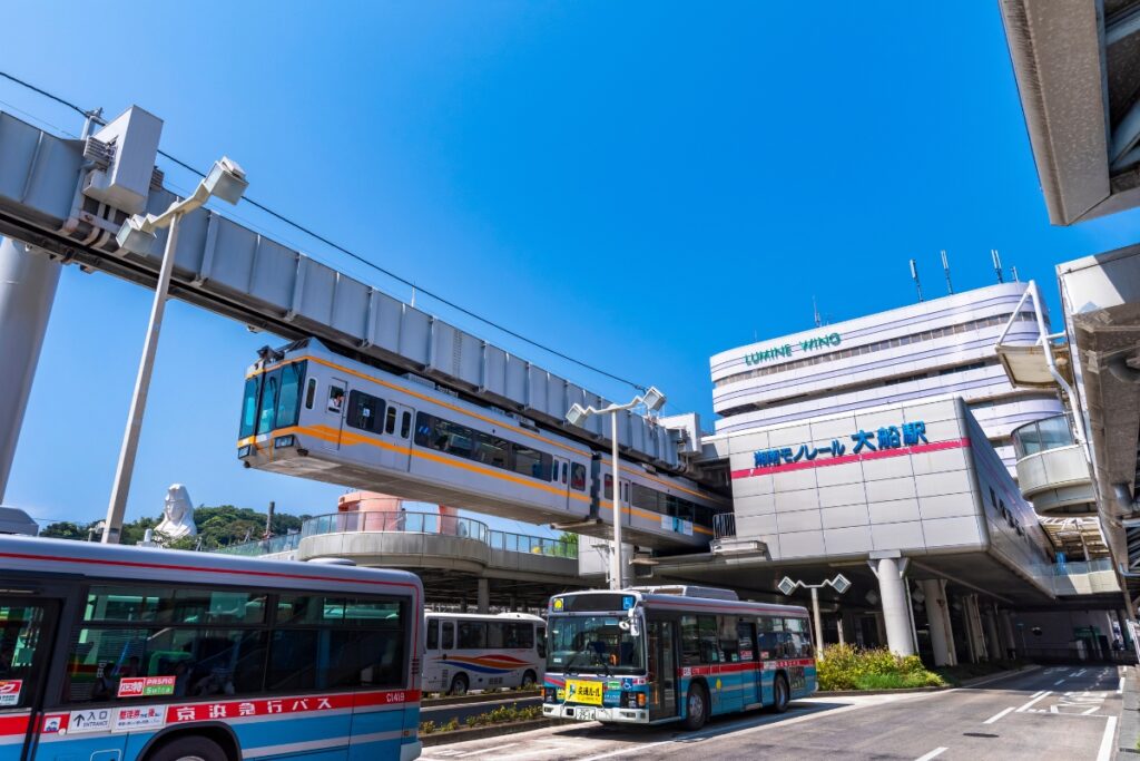A Shonan Monorail train departs from Ofuna Station on an elevated track, with buses parked below and the station building in the background. The sky is clear and blue.