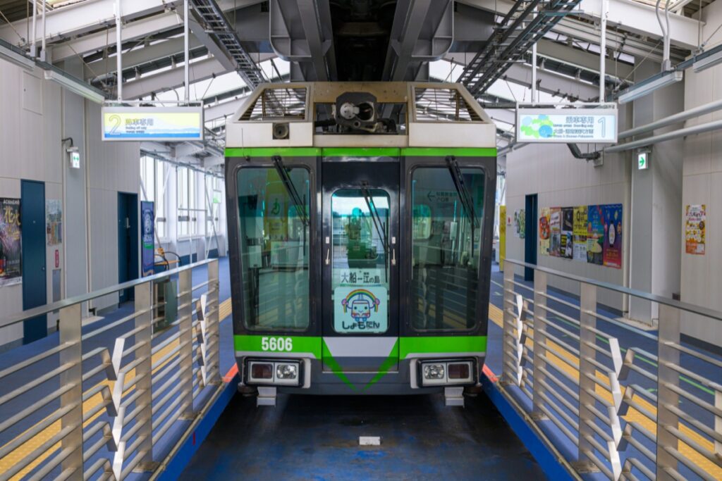 A Shonan Monorail train with green accents, labeled "5606," is stopped at the Ofuna station platform. The indoor station has signs, railings, and advertisements on the walls.