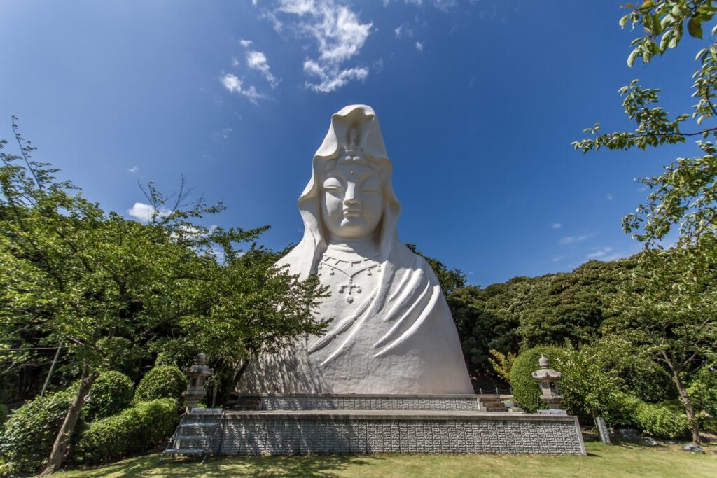 A large white statue of Kannon, the Buddhist goddess of mercy, surrounded by trees and greenery under a clear blue sky. The statue features intricate details and sits on a stone platform.