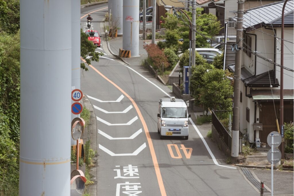 A narrow suburban street with vehicles, including a small white van and a red car, passing under large monorail support columns. The street is lined with greenery, houses, and utility poles.