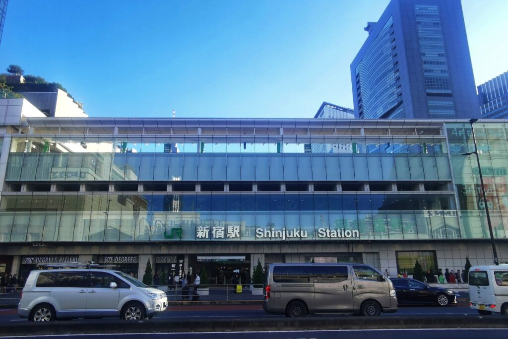 The exterior of Shinjuku Station in Tokyo, Japan, with its modern glass facade reflecting the blue sky and surrounding skyscrapers. The station's signage in both Japanese and English is prominently displayed above the entrance. In the foreground, several vehicles, including a silver van, are seen on the busy street, while pedestrians walk along the sidewalk.