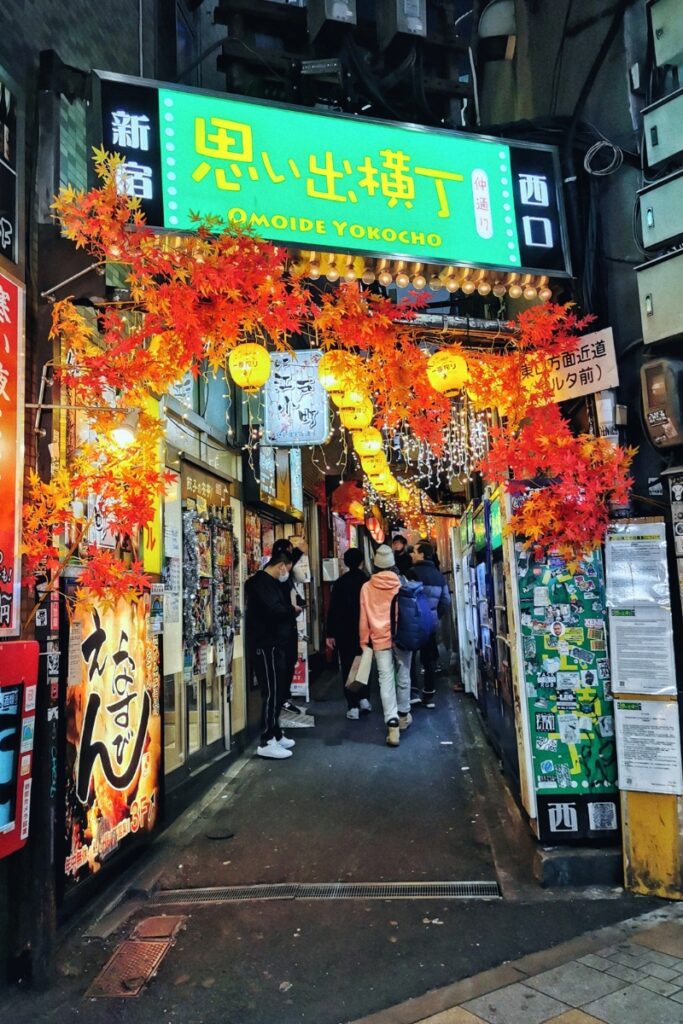 A vibrant entrance to Omoide Yokocho, a narrow alley in Shinjuku, Tokyo, lined with small restaurants and izakayas. The green neon sign with Japanese characters and 'OMOIDE YOKOCHO' in English glows above, surrounded by warm yellow lanterns and bright red autumn leaves. People walk through the bustling alley, passing by colorful posters, menus, and sticker-covered walls, adding to the lively atmosphere of this iconic dining street.