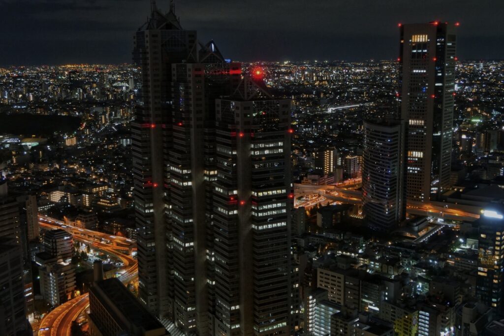 A breathtaking night view of Shinjuku and the greater Tokyo area, illuminated by countless city lights. Skyscrapers with glowing windows and red aviation lights stand tall against the dark sky, while highways weave through the urban landscape with golden streams of traffic. The city's vast expanse stretches toward the horizon, showcasing Tokyo's vibrant nighttime energy.