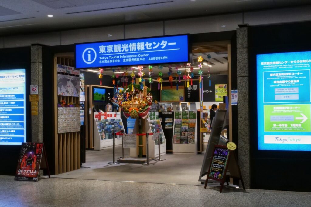 A well-lit Tokyo Tourist Information Center entrance with a bright blue sign in Japanese and English. Inside, brochures, maps, and travel guides are neatly displayed, while colorful decorations, including a festive ornament, hang from the ceiling. Digital screens on either side provide additional tourist information, and a 'Godzilla: Attack on Tokyo' sign stands near the entrance, adding a touch of pop culture.