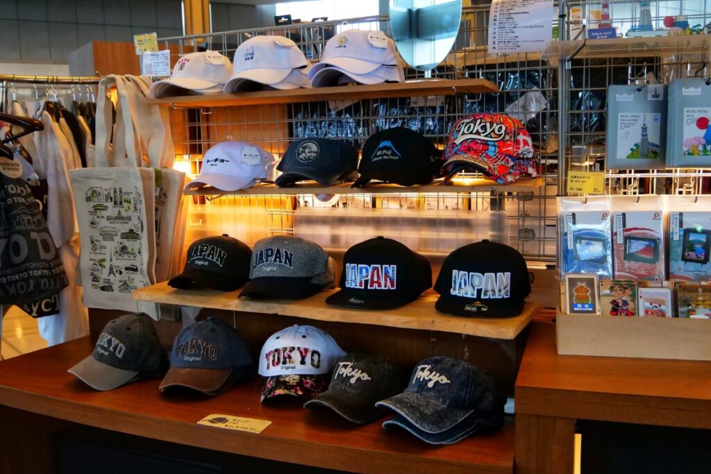 A display of souvenir caps and tote bags at a gift shop, featuring various designs with 'Tokyo' and 'Japan' embroidered on them. The hats come in different colors and styles, including baseball caps and snapbacks. Nearby shelves also hold postcards and other memorabilia, creating a well-stocked souvenir section.