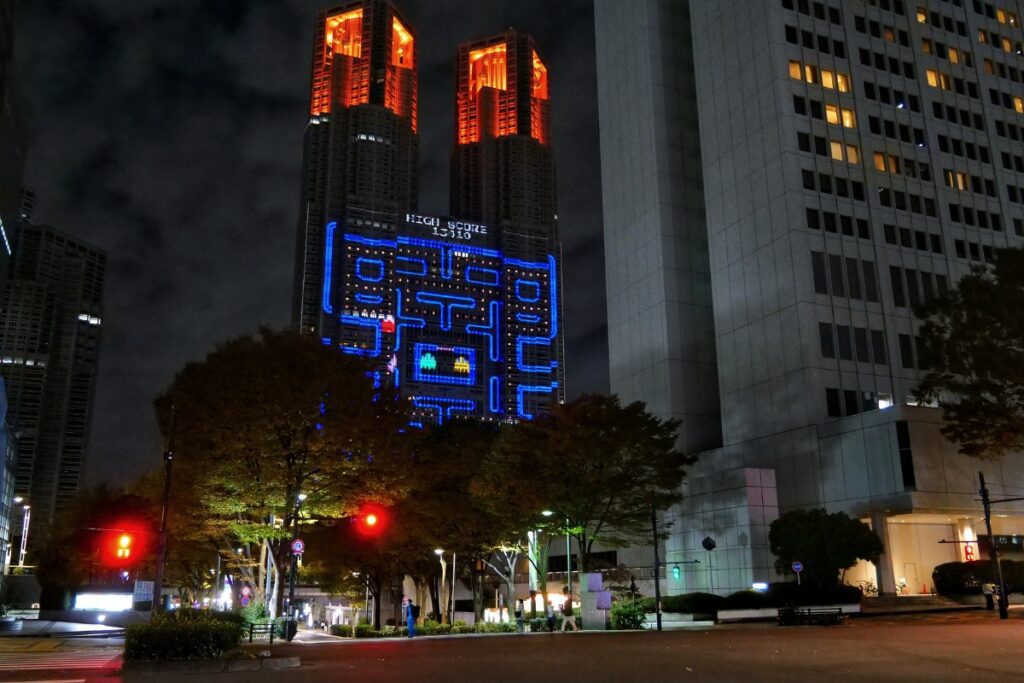 The Tokyo Metropolitan Government Building illuminated at night with a massive projection of a Pac-Man game on its facade. The game's neon blue maze and bright yellow Pac-Man stand out against the dark skyscraper, while the building's upper sections glow with orange lights. Trees and a traffic signal with a red light are visible in the foreground, adding depth to the urban scene.