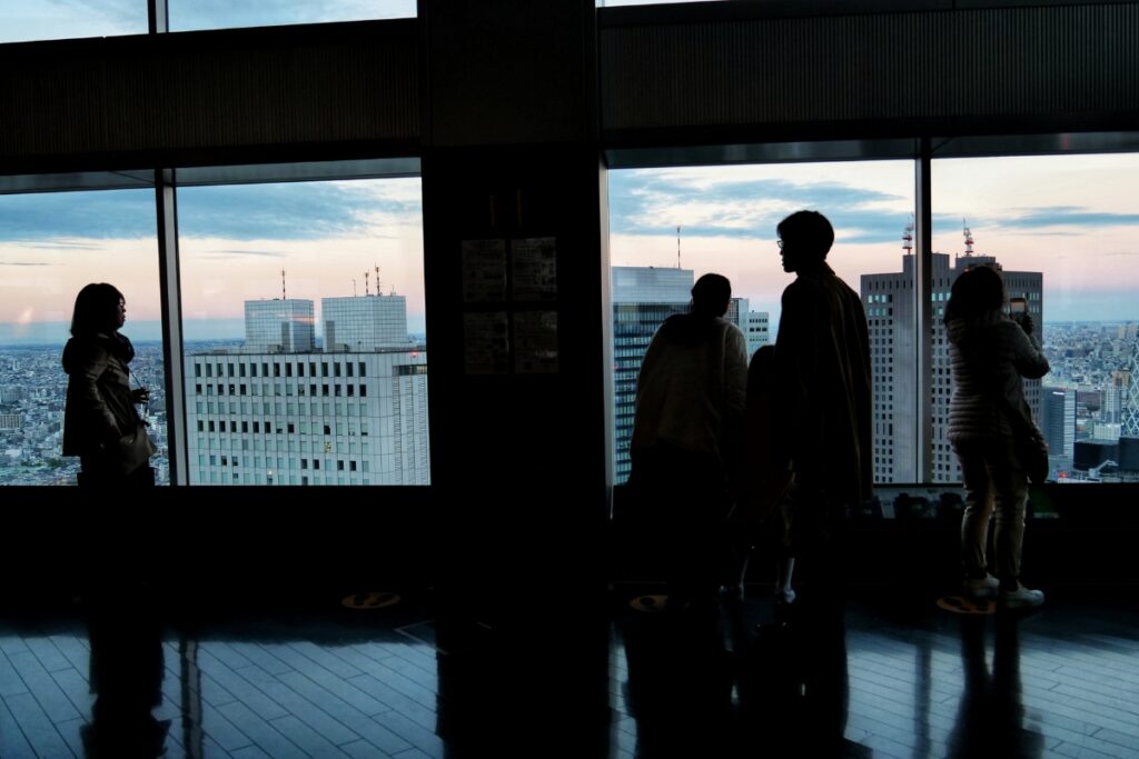 Silhouetted visitors stand by large windows, admiring a panoramic view of Shinjuku City at dusk. The skyline features modern skyscrapers with antennas, while the sky transitions from soft pink to deep blue. Reflections of the onlookers on the polished floor add depth to the serene atmosphere of this high-rise observation deck