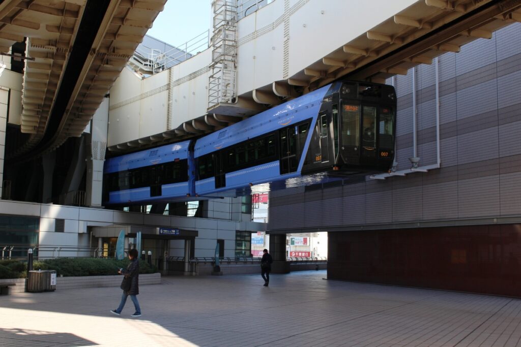A sleek blue Chiba Monorail train is seen gliding through an urban station, suspended from an overhead track. The modern train features a streamlined black front with digital signage displaying its route. Below, pedestrians walk across a spacious tiled plaza, with one person wearing a dark coat and jeans carrying a phone. The station architecture combines glass, metal, and tiled surfaces, with railings and signage visible in the background. Sunlight casts long shadows on the ground, highlighting the train’s elevated and futuristic design.