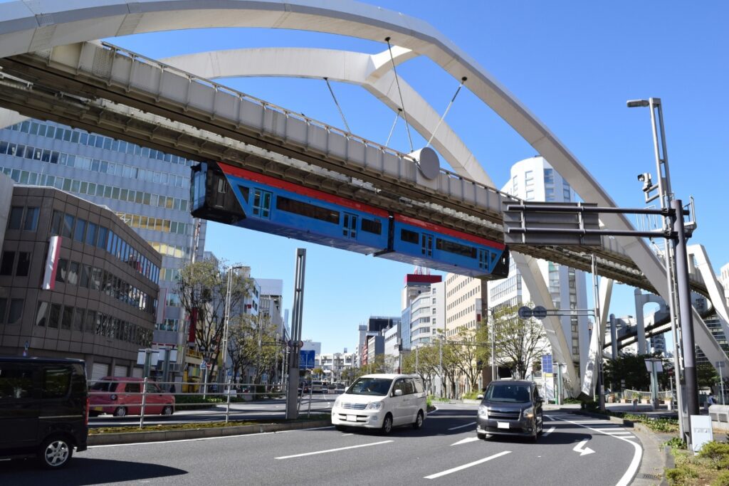 A blue and red suspended monorail train travels along an elevated track supported by white arched structures in an urban setting. Below, cars and vans navigate a curved road surrounded by modern buildings with large glass windows. The sky is clear and blue, and trees line the sidewalks, adding greenery to the scene. The monorail, part of a hanging railway system, appears to be moving above the road, creating a striking contrast against the cityscape.