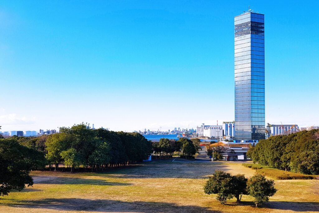 A tall, sleek, glass-covered skyscraper, Chiba Port Tower, rises against a clear blue sky in the background. The foreground features a spacious park with a mix of green and dry grass, scattered trees, and long shadows cast by the sunlight. Beyond the park, an industrial harbor area with large storage tanks and cranes is visible, with the distant Tokyo skyline faintly appearing on the horizon. The deep blue water of the bay contrasts with the greenery and urban structures, creating a balanced composition of nature and modern architecture.