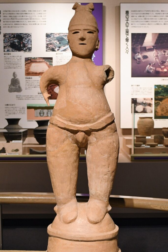 A Haniwa clay statue of a sumo wrestler displayed in a museum. The ancient terracotta figure has a rounded body, a traditional sumo-style belt, and a headdress. The statueโs arms are broken off, leaving only partial stumps. It stands on a cylindrical base, characteristic of Haniwa funerary sculptures from Japanโs Kofun period. The face has simple, hollowed-out eyes and a neutral expression. In the background, museum display panels feature historical information, images of archaeological sites, and artifacts related to ancient Japanese culture.