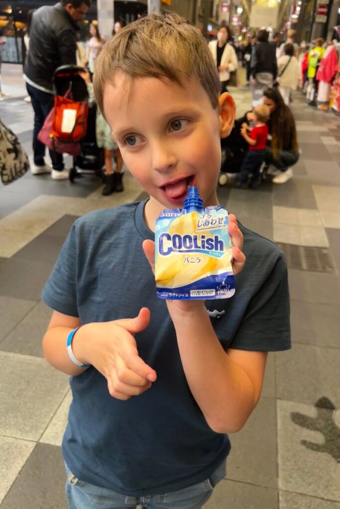 A young boy with short brown hair and a playful expression enjoys a Coolish vanilla ice cream pouch, licking the spout while holding it up to the camera. He wears a dark blue t-shirt and a blue wristband on his left wrist. The background shows a lively shopping arcade with people walking, shopping, and pushing strollers, creating a bustling atmosphere.