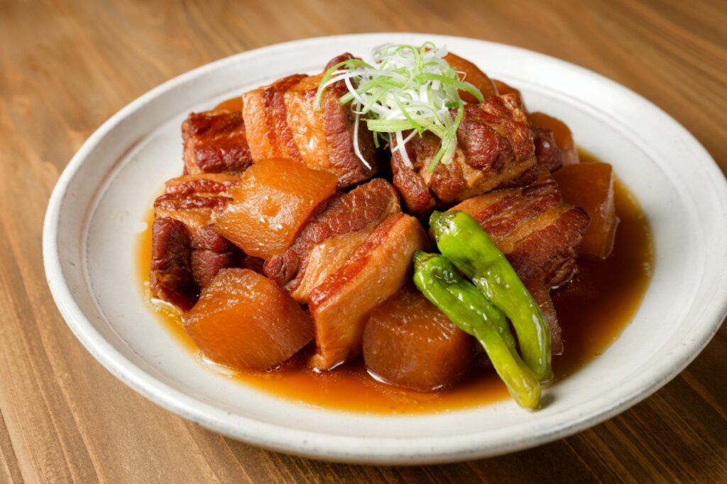 A plate of kakuni, Japanese braised pork belly, served with simmered daikon radish and garnished with thinly sliced green onions. The pork is caramelized and glistening with a rich soy-based sauce, accompanied by bright green shishito peppers. The dish is presented on a white ceramic plate, set on a wooden table.