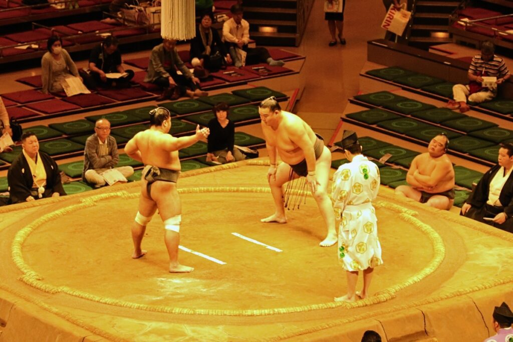 Two sumo wrestlers face off in a traditional sumo ring (dohyō) during a tournament. One wrestler wears a black mawashi (sumo belt) with white sagari (decorative strings), while the other also wears a black mawashi. The wrestlers are in a pre-bout stance, crouching and preparing for the match. A gyōji (referee) dressed in a patterned white and green kimono stands between them. Around the ring, seated spectators watch attentively, with some sitting on traditional floor cushions. In the background, other sumo wrestlers and officials observe the bout.