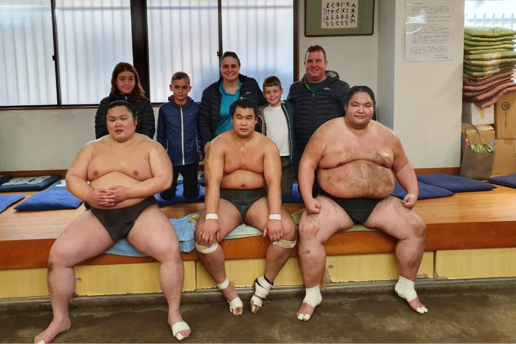 A group of sumo wrestlers from the Ogurama Stable sits on a wooden bench in their traditional training attire after a practice session, with their feet and wrists wrapped in protective tape. Behind them, a smiling family of five, dressed in jackets, poses for a photo inside the sumo training facility. The background includes stacked cushions, framed calligraphy, and training equipment, adding to the authentic atmosphere of the sumo stable.