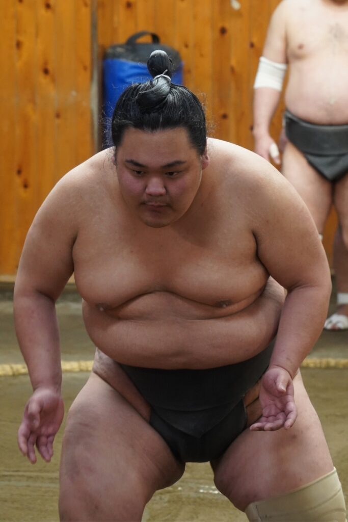 A sumo wrestler with a focused expression is crouched in a ready stance during training. He wears a black mawashi (sumo belt) and has his hair styled in a traditional chonmage (topknot). His legs are wrapped with bandages for support. In the background, another sumo wrestler in a similar outfit is visible, along with a wooden training area featuring walls and a padded floor. The wrestler's intense gaze and poised stance reflect concentration and discipline.