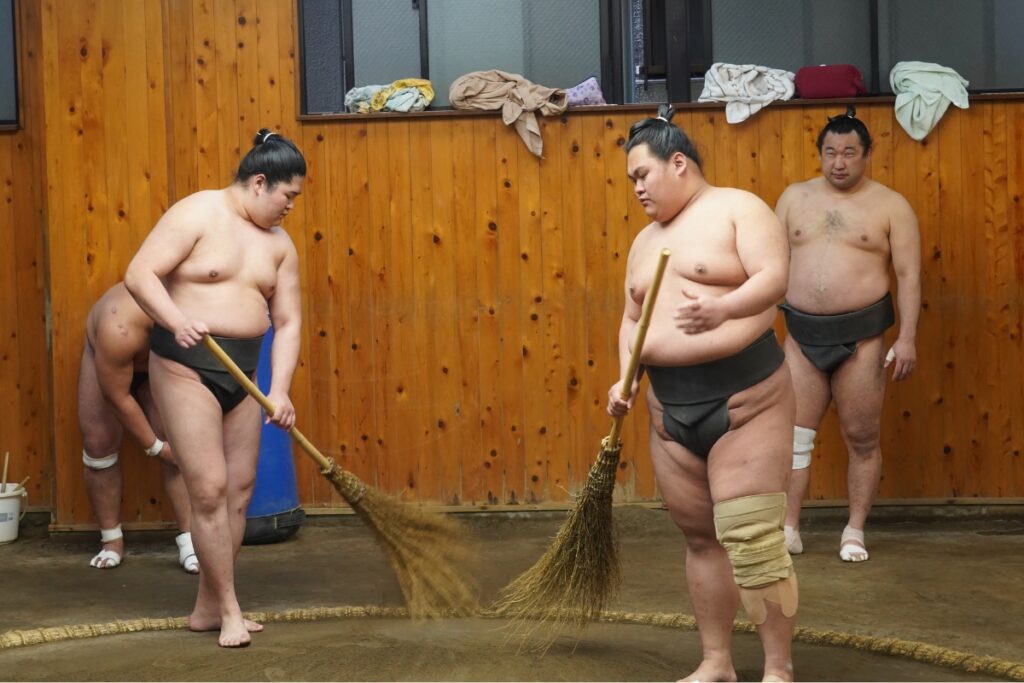 Sumo wrestlers in a training stable clean the ring using traditional brooms made of straw. They are dressed in black mawashi (sumo belts), with one wrestler having his knee wrapped in a support bandage. The wooden walls in the background have towels and clothes draped over the ledge, and a bucket with cleaning tools is visible in the corner. Another wrestler stands in the background, observing the cleaning process.