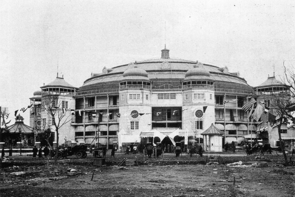 Black and white photograph of the original Ryogoku Kokugikan sumo arena in Tokyo, taken in 1909. The grand circular structure features multiple domed towers, balconies, and arched windows, showcasing an architectural style blending Western and Japanese influences. Flags and banners decorate the building's facade, indicating a special event or grand opening. In the foreground, people in early 20th-century attire walk along a dirt path, while horse-drawn carriages and an early automobile are visible near the entrance. The surrounding area appears underdeveloped, with scattered wooden debris and bare trees, highlighting the contrast between the newly built arena and its surroundings.