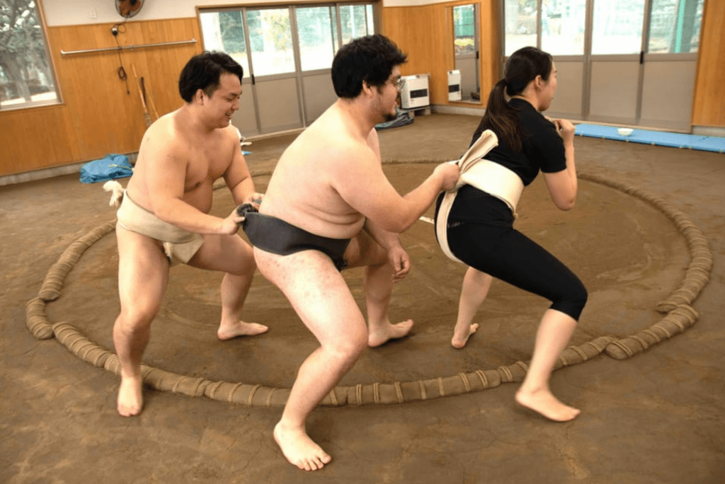 Two sumo wrestlers in traditional mawashi belts and a woman in athletic wear are engaged in a playful training drill inside a sumo ring. Each person is holding the belt of the person in front, forming a humorous training chain, suggesting a hands-on sumo wrestling experience at the Amita Sumo training facility in Tokyo.