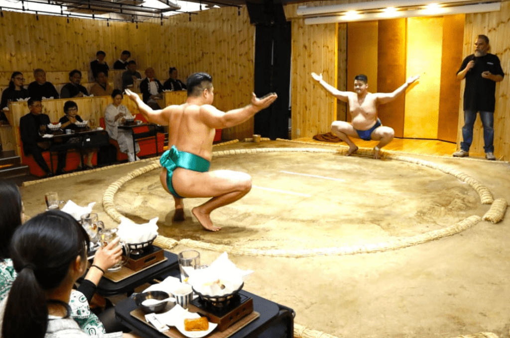 Two sumo wrestlers wearing colorful mawashi face off in a ceremonial stance inside a small sumo ring at the Askasua Sumo Club Show in Tokyo, while an announcer stands to the side. The audience, seated at tables with traditional Japanese meals, watches the live sumo demonstration in an intimate wooden venue, blending sport with dining entertainment.