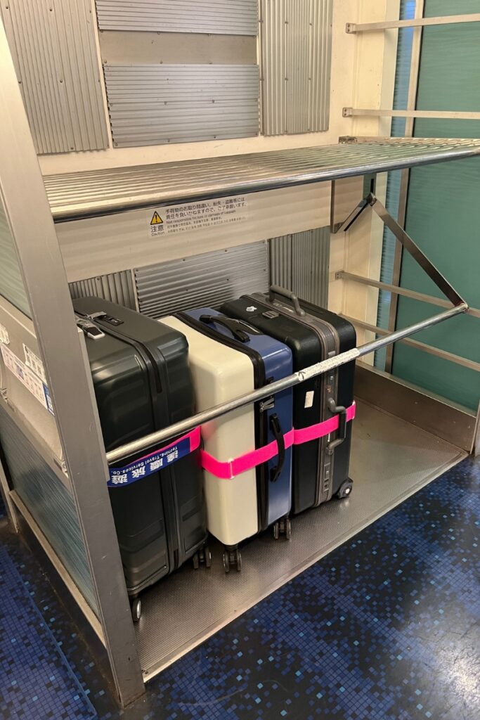 A designated luggage storage area at the end of a Keisei Skyliner train car, featuring three large suitcases secured with a bright pink strap. The storage rack has a metal frame with horizontal bars to hold baggage in place. A warning sign in Japanese is visible, advising passengers to secure their luggage properly. The train's interior includes blue patterned flooring and metallic wall panels, reflecting a sleek and modern design.