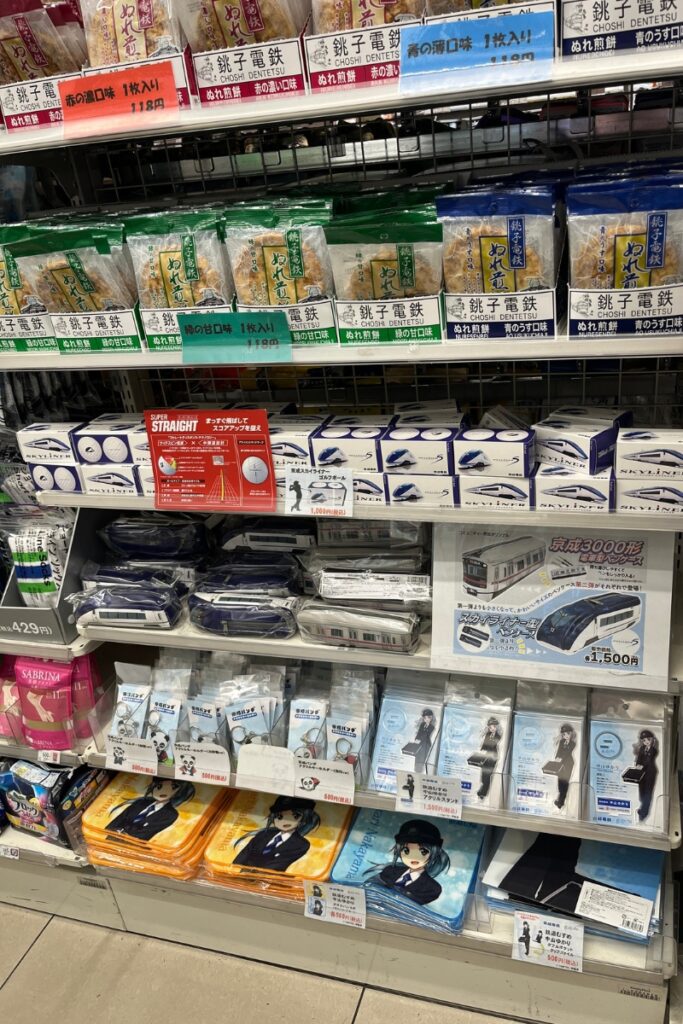 A store shelf stocked with Keisei Skyliner-themed merchandise and local snacks. The upper shelves contain various packaged snacks, including Choshi Dentetsu-branded senbei rice crackers in green, blue, and red packaging. Below, an array of Skyliner souvenirs is displayed, including train-themed model kits, pencil cases, and keychains featuring the high-speed train’s design. A red promotional sign highlights a specific product. The lower section features anime-style character illustrations on hand towels and other collectibles, depicting a female train conductor in uniform. The neatly arranged items cater to train enthusiasts and travelers looking for themed memorabilia.